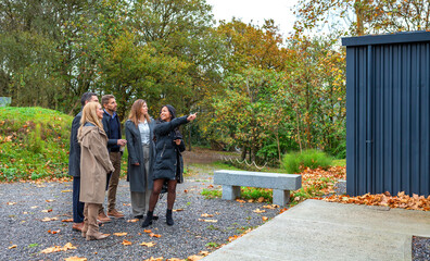 Female real estate agent pointing to home built with shipping containers while presenting the property to investors in an autumn visit. Contemporary sustainable and eco friendly architecture concept.