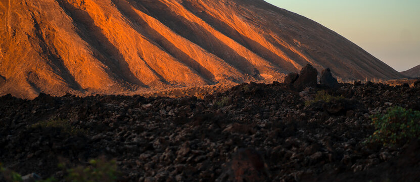 A volcanic landscape with black lava formations and a mountain in the sun. - Powered by Adobe
