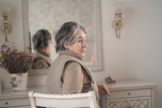 Beautiful old woman sits near the mirror in her cozy light room.