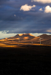 A wind farm with windmills among the desert rocky land and mountains at sunset.