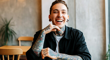 Smiling young man with tattoos, wearing a black shirt, sitting at a wooden table in a cozy cafe, showcasing a joyful expression and relaxed atmosphere