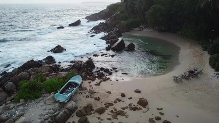 Aerial view of ocean waves breaking against rocky coastline, surrounding palm fringed landscape with aged boat drifting near sandy shore