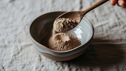 Hand pouring fine brown powder from a wooden spoon into a rustic ceramic bowl filled with more powder, creating a small crater.