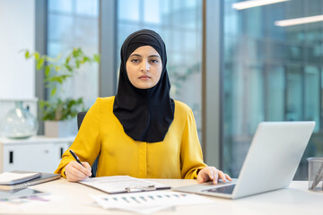 Muslim businesswoman wearing a hijab, looking directly at the camera while working on a laptop and writing notes at her desk in a modern corporate office, representing dedication and professionalism