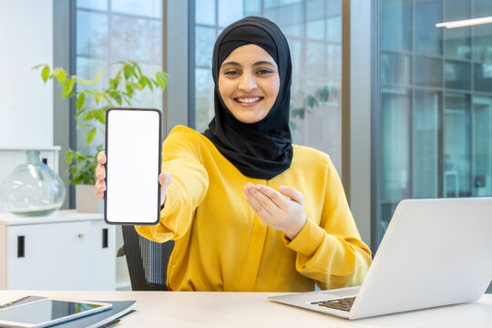 Muslim business woman wearing a hijab showing a blank white smartphone screen mockup while smiling and gesturing at a modern office desk, with a laptop and tablet - Powered by Adobe