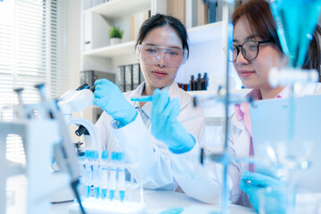 Two female scientists in white lab coats working together in a bright laboratory with microscopes, glassware, and blue test tubes, representing teamwork and modern scientific research environment.