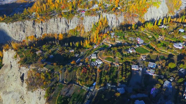 Aerial view of houses amidst the colorful trees and terraced fields, creating a vibrant tapestry against the backdrop of towering cliffs, Hunza, Gilgit Baltistan, Pakistan.