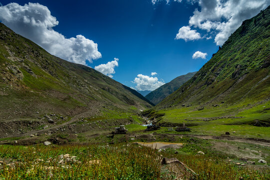 View of a lush green valley with steep mountainsides under a bright blue sky dotted with fluffy clouds, a small stream trickling through, Noori Top, Khyber Pakhtunkhwa, Pakistan.