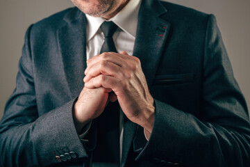 Businessman in dark suit with hands firmly clasped together in front of his chest, conveying intense worry, hope, prayer, or desperation