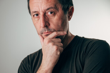Portrait of a middle-aged man with short dark hair and stubble, gazing thoughtfully with hand on chin against neutral gray background. Reflection, worry and decision-making concept.