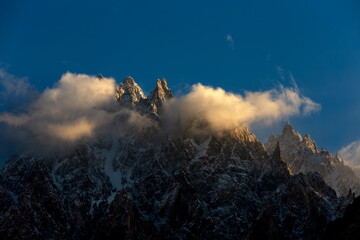 View of jagged, snow-dusted peaks piercing through a veil of luminous clouds under a clear sky, Karakoram Highway, Passu, Gilgit Baltistan, Pakistan.