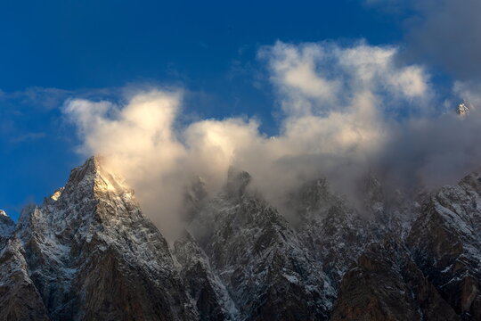 View of jagged, snow-dusted peaks piercing the azure sky, partially veiled by ethereal clouds, creating a majestic dance of light and shadow, Passu, Gilgit Baltistan, Pakistan.