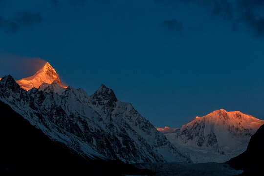 View of stark, snow-capped peaks kissed by the golden light of the setting sun against a deep blue sky, Karakoram Highway, Passu, Gilgit Baltistan, Pakistan.