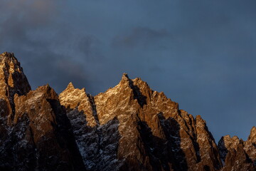 View of jagged, dark mountains kissed by golden sunlight under a moody blue sky, casting long shadows on the rugged terrain, Passu, Gilgit Baltistan, Pakistan.