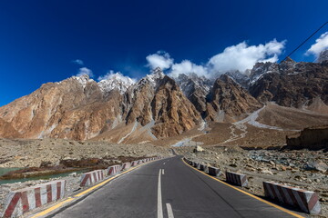 View of stark grey road contrasting with rugged, snow-dusted peaks piercing the azure sky, framed by rocky terrain and wispy clouds, Passu, Gilgit Baltistan, Pakistan.