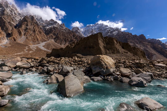 View of turquoise river rushing past massive boulders under jagged, snow-dusted peaks against a clear blue sky with clouds, Passu, Gilgit Baltistan, Pakistan.