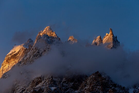 View of golden sunlight kissing the snow-dusted peaks rising majestically above the swirling clouds along the Karakoram Highway, Passu, Gilgit Baltistan, Pakistan.