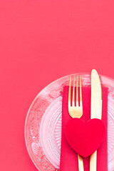 A close-up of a place setting for a romantic dinner, featuring a clear glass plate, gold cutlery, and a red velvet heart on a red napkin, all on a bright red background.