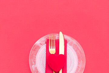 romantic dinner, featuring a clear glass plate, gold cutlery, and a red velvet heart on a red napkin, all on a bright red background. The shot is cropped tightly to emphasize the details.