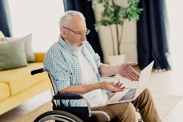 Elderly man in wheelchair uses laptop at home in living room, exploring online learning and communication opportunities