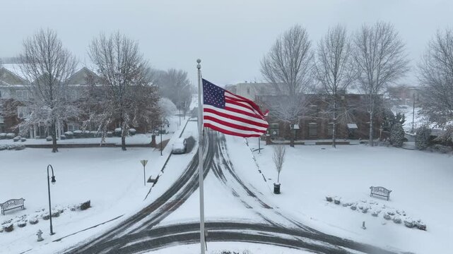 American flag on flagpole in center of snowy roundabout. Aerial approaching shot. Snowy winter snowfall in american town. Empty roads and apartment houses in background.