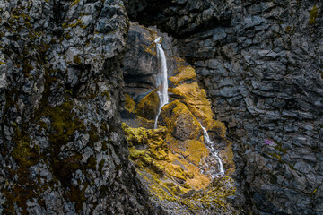 Aerial view of a serene waterfall cascading down the rugged cliffs, moss-covered rocks adding a touch of vibrant color, Seljavallalaug, Rangárþing eystra, Iceland.