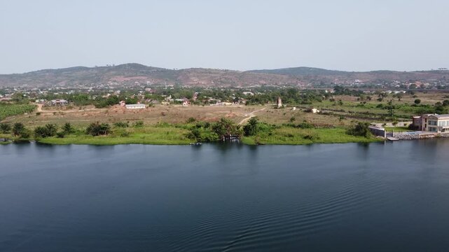 Aerial drone footage of Ghana, Africa, serene lake surrounded by mountains. Traditional boats across the water near a local village, with views near the Right to Dream Football Academy