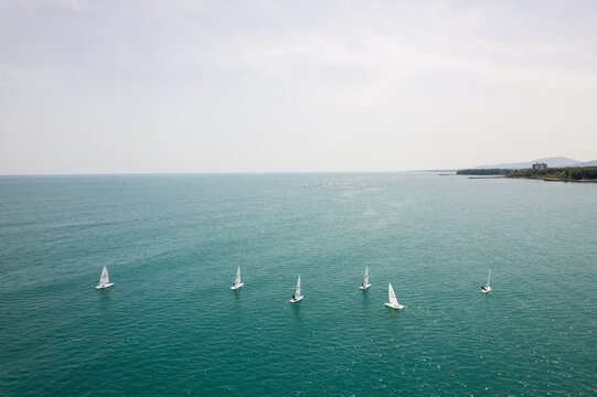 Aerial view of sailboats scattered like delicate paper boats on a vast expanse of turquoise water, contrasting with the distant, hazy coastline, Primorsko, Burgas, Bulgaria.
