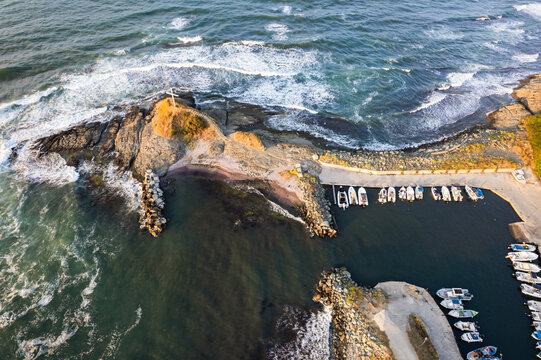 Aerial view of waves crashing against the rugged coastline near a small port filled with boats, a serene blend of nature and human presence, Lozenets Southern Cross, Lozenets, Tsarevo, Bulgaria.