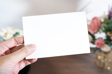 Obraz premium Mockup of a blank white card held by hand with soft blurred flowers, perfect for wedding or Valentine’s greeting design, with clipping path.