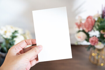 Obraz premium Mockup of a blank white card held by hand with soft blurred flowers, perfect for wedding or Valentine’s greeting design, with clipping path.
