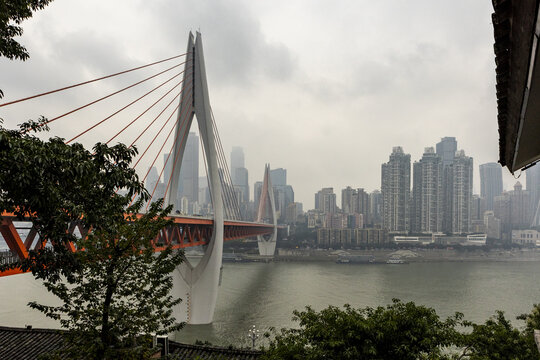 View of a striking orange bridge contrasts against the modern skyline and tranquil waters on an overcast day, Chongqing, Chongqing, China.
