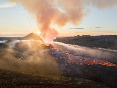 Aerial view of a fiery volcanic eruption spewing smoke and lava against a backdrop of green fields and a radiant sunset, Fagradalsfjall Volcano, Grindav&iacute;kurbaer, Iceland.