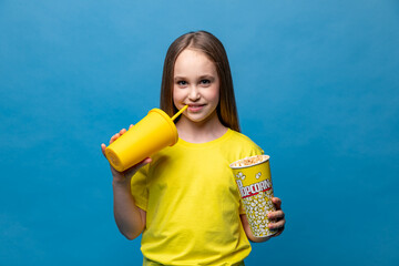 Kids cinema deals. Happy little girl wearing yellow t-shirt, watch movie film, holds bucket of popcorn cup and yellow plastic glass with beverage on yellow background, copy space