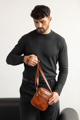 A stylish man in a dark grey crew neck sweater poses with a cognac leather crossbody bag against a minimalist white studio background.