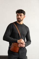 A stylish man in a dark grey crew neck sweater poses with a cognac leather crossbody bag against a minimalist white studio background.