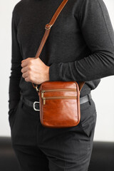 Male model in a charcoal knit crewneck and tailored trousers wearing a cognac leather vertical messenger bag against a white studio background.