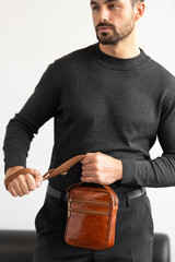 A stylish man in a dark grey crew neck sweater poses with a cognac leather crossbody bag against a minimalist white studio background.