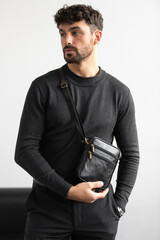 A stylish man in a dark grey crew neck sweater poses with a black leather crossbody bag against a minimalist white studio background.