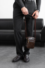 Stylish man in charcoal knitwear and trousers holds a dark brown vertical leather messenger bag against a white studio backdrop