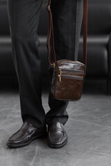Stylish man in charcoal knitwear and trousers holds a dark brown vertical leather messenger bag against a white studio backdrop