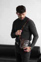 Stylish man in charcoal knitwear and trousers holds a dark brown vertical leather messenger bag against a white studio backdrop