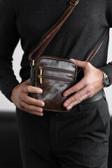 Stylish man in charcoal knitwear and trousers holds a dark brown vertical leather messenger bag against a white studio backdrop