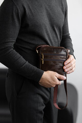 Stylish man in charcoal knitwear and trousers holds a dark brown vertical leather messenger bag against a white studio backdrop