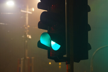 A green traffic signal glowing through dense urban fog during nighttime. Illustrates reduced visibility conditions, road safety management, smart city infrastructure, and climate-affected mobility.
