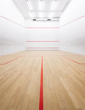 Indoor squash court with wooden floor and white walls. Symmetrical composition - wide-angle perspective view of well-lit squash court with red markings.