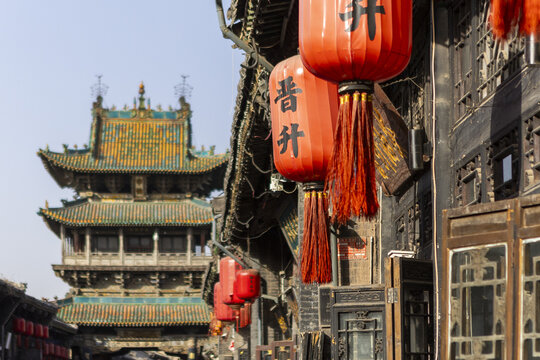 View of vibrant red lanterns hanging against aged dark wooden structures, leading the eye towards a distant, ornate tower, Pingyao, Shanxi, China.
