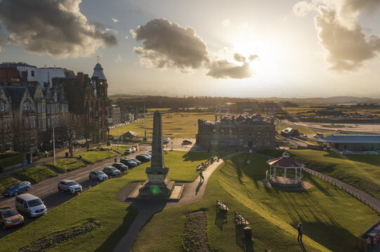 Aerial view of a sun-kissed obelisk standing tall amidst lush greenery and the iconic Old Course, casting shadows under a dramatic sky, St Andrews, Scotland, United Kingdom.