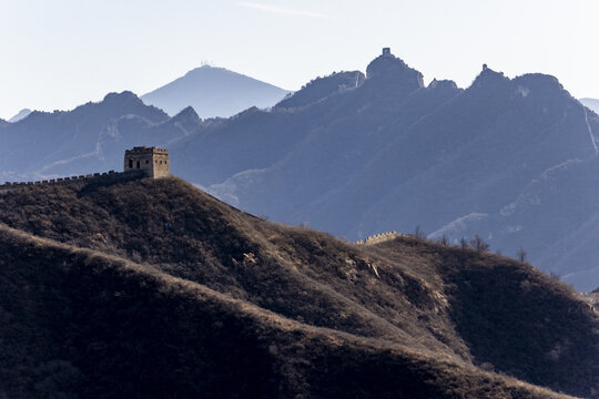 View of the ancient Great Wall snaking across rugged, sun-kissed hills under a hazy sky, a testament to history and human endeavor, Chengde, Beijing, China.