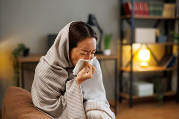 Sick Asian woman blowing nose with tissue wrapped in warm blanket